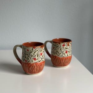 May include: Two ceramic mugs with a floral design. The mugs have a cream-colored background with green leaves and red flowers. The bottom half of the mugs is a textured orange-brown color. Each mug has a gray handle and a brown interior.
