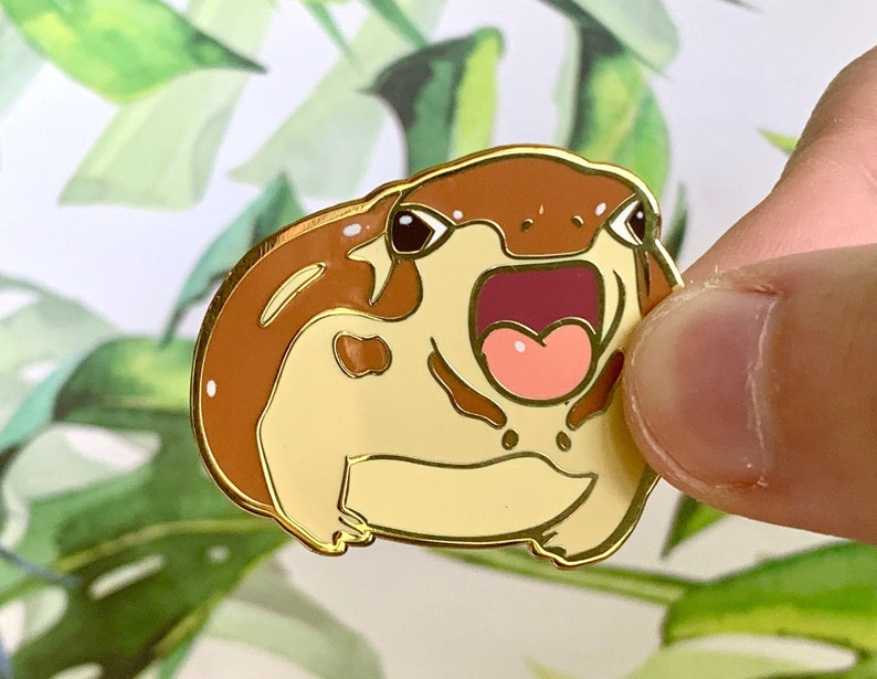 May include: A gold-tone enamel pin of a cartoon frog with a brown back, yellow belly, and a wide open mouth. The frog has black eyes and a pink tongue.