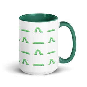 May include: White ceramic mug with a green rim and handle. The mug features a repeating pattern of green caterpillars in different positions.
