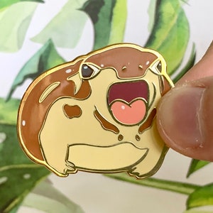 May include: A gold-colored enamel pin of a cartoon frog with brown and beige markings. The frog has its mouth open wide, revealing a pink tongue and teeth.