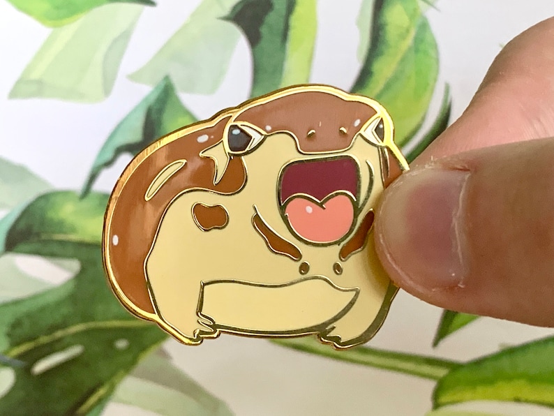 May include: A gold-colored enamel pin of a cartoon frog with brown and beige markings. The frog has its mouth open wide, revealing a pink tongue and teeth.