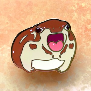 May include: A brown and white enamel pin of a frog with its mouth open wide, showing its pink tongue. The frog has brown spots on its body and black eyes.