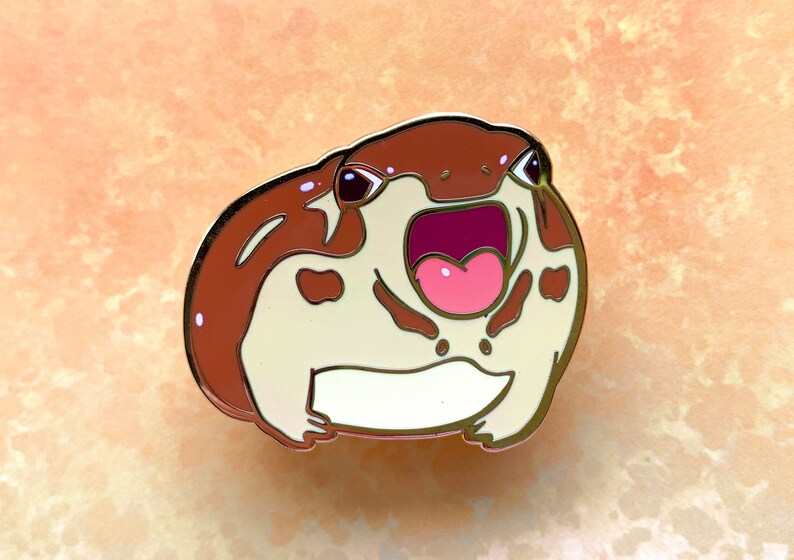May include: A brown and white enamel pin of a frog with its mouth open wide, showing its pink tongue. The frog has brown spots on its body and black eyes.
