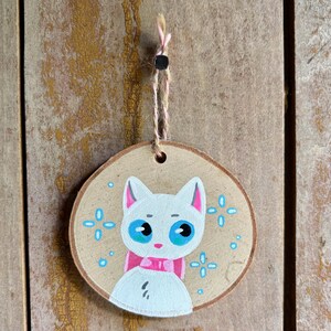 Puede incluir: Un adorno redondo de madera con un gato blanco pintado a mano, con grandes ojos azules, orejas rosadas y una pajarita rosa. El gato está rodeado de pequeños detalles en forma de estrella azul claro. Un cordel fino está sujeto en la parte superior.