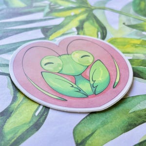 May include: A green praying mantis with large eyes is depicted inside a pink heart. The sticker is perfect for adding a touch of whimsy to any surface.