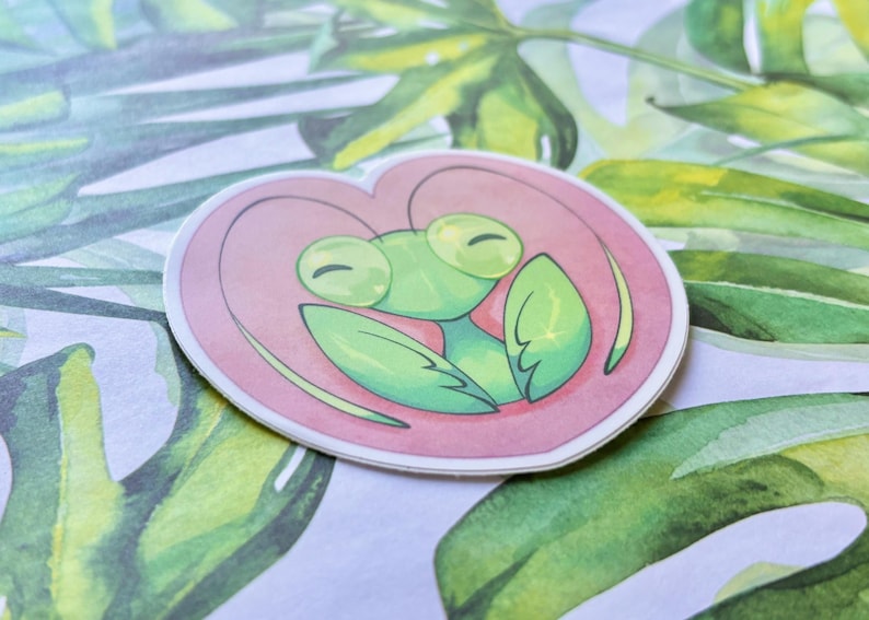 May include: A green praying mantis with large eyes is depicted inside a pink heart. The sticker is perfect for adding a touch of whimsy to any surface.