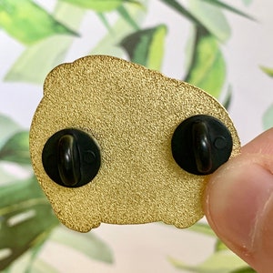 May include: Gold-colored enamel pin with two black metal clasps on the back. The pin is shaped like a cartoon character.