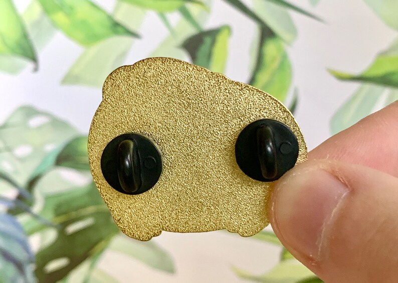 May include: Gold-colored enamel pin with two black metal clasps on the back. The pin is shaped like a cartoon character.