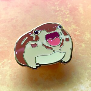 May include: A brown and white enamel pin of a frog with its mouth open and a pink tongue. The frog has brown spots on its body.