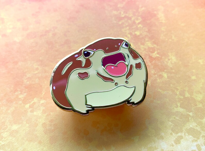 May include: A brown and white enamel pin of a frog with its mouth open and a pink tongue. The frog has brown spots on its body.