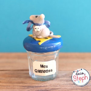 May include: A small glass jar with a blue lid and a decorative mouse figurine on top. The mouse is gray with a blue ribbon and a small tag. The jar has a label that reads "Mes Quenottes".