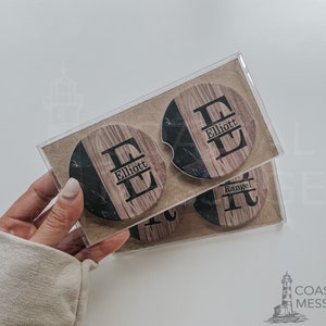 May include: Set of four personalized car coasters. Each coaster features a wood grain and black marble design with a large initial and a last name. The coasters are packaged in a clear plastic box.