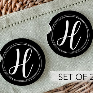 Può includere: Set di due sottobicchieri neri per auto con la lettera "H" bianca all'interno di un design circolare bianco. I sottobicchieri hanno un intaglio curvo. Il testo "SET OF 2" è visualizzato in bianco su sfondo rettangolare bianco.