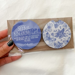 May include: Two car coasters. One coaster is blue with the text "SEEK the KINGDOM of GOD" and a Bible verse. The other coaster has a blue floral and butterfly pattern. Both are packaged together.