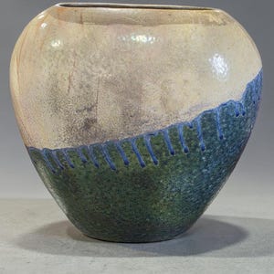 May include: A handcrafted ceramic vase with a rounded shape. The top half is a light beige color, transitioning to a green and blue textured bottom half. Blue lines decorate the border between the colors. A unique piece for home decor.