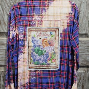 May include: A blue and pink plaid flannel shirt with a bleached effect. The shirt has a patch on the back with a fairy and flowers and the text "Lilac Cottage Fairy".