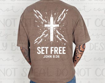 May include: Brown t-shirt with a white cross design, chains, and the text "SET FREE" and "JOHN 8:36". The shirt is a neutral color and has short sleeves.