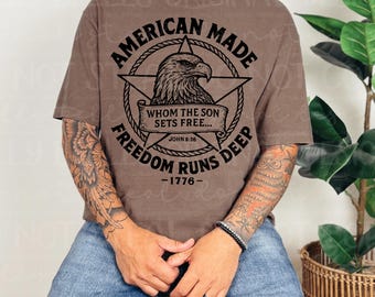 May include: Brown t-shirt with the text "AMERICAN MADE" in a circular design, featuring an eagle and the words "FREEDOM RUNS DEEP - 1776". The shirt also includes the Bible verse John 8:36. The shirt is a neutral color.
