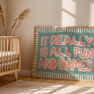 Puede incluir: Una impresión artística enmarcada con el texto "IT REALLY IS ALL FUN AND GAMES" en letras coral sobre un fondo verde azulado. La obra de arte está en un marco de madera clara y se exhibe en un entorno de guardería con una cuna de madera.