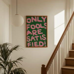 May include: A framed art print with the text "ONLY FOOLS ARE SATISFIED!" in pink letters on a green background. The print is in a brown wooden frame and hangs on a white wall. A staircase and a hanging light are also visible.