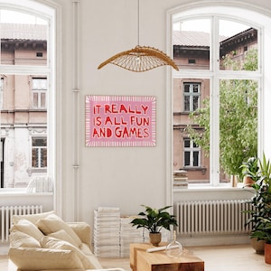 May include: A framed pink and white art print with the text "IT REALLY IS ALL FUN AND GAMES" hangs on a white wall. The room features a beige sofa, wooden coffee table, and a woven pendant light. Large windows offer a view of an urban setting.
