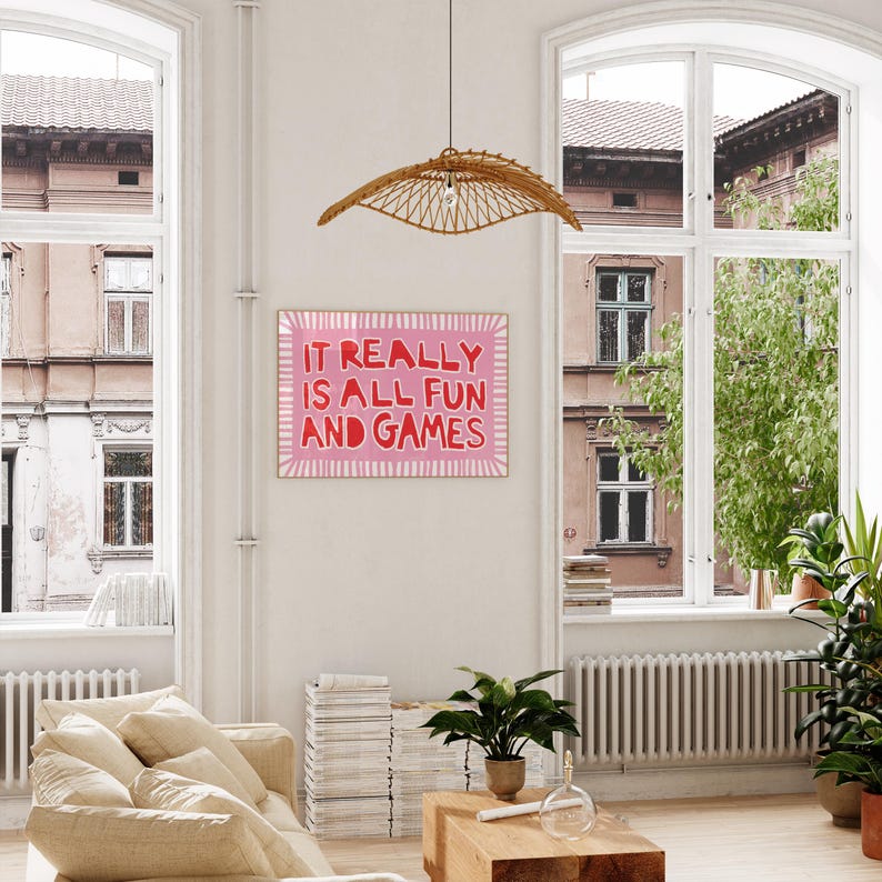 May include: A framed pink and white art print with the text "IT REALLY IS ALL FUN AND GAMES" hangs on a white wall. The room features a beige sofa, wooden coffee table, and a woven pendant light. Large windows offer a view of an urban setting.