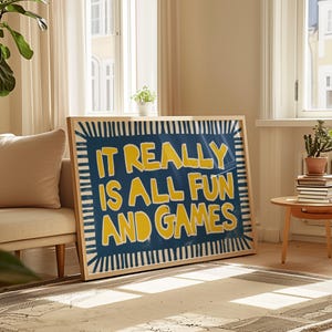 May include: A framed art print with the text "IT REALLY IS ALL FUN AND GAMES" in yellow on a blue background. The frame is light-colored wood. The artwork is leaning against a wall in a living room setting.