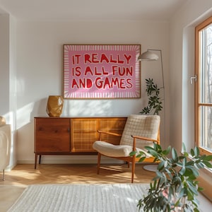 Framed typography wall art reading “It Really Is All Fun and Games” in red lettering on a pink background, styled in a sunlit living room with a mid-century wood console, lounge chair, floor lamp, and plants.