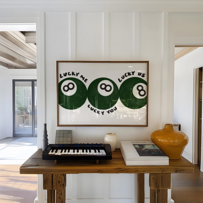 Puede incluir: Una obra de arte enmarcada con tres dise&ntilde;os de bola 8 verdes con el texto "LUCKY ME", "LUCKY YOU" y "LUCKY US". La obra cuelga sobre una consola de madera con un teclado peque&ntilde;o, un jarr&oacute;n blanco y un jarr&oacute;n decorativo amarillo-naranja.