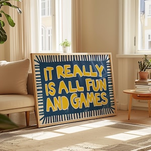 May include: A framed art print with the text "IT REALLY IS ALL FUN AND GAMES" in yellow on a blue background. The artwork is in a light wooden frame and rests on a rug in a living room setting.