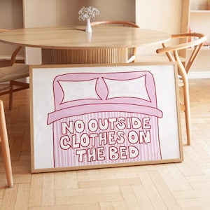 May include: A framed art print featuring a pink bed illustration and the text "NO OUTSIDE CLOTHES ON THE BED". The artwork has a white background with pink stripes and a wooden frame. The print is displayed in a room with wooden furniture.