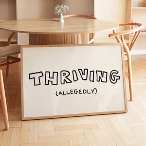 以下が含まれることがあります： 遊び心のある手書き風の黒い文字で「THRIVING (ALLEGEDLY)」と書かれた白い額装のプリント。プリントは明るい木製の床のある部屋の木製のテーブルに立てかけられています。