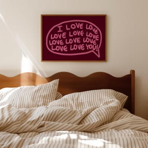 May include: A framed print with the words "I LOVE LOVE LOVE LOVE LOVE LOVE LOVE LOVE YOU!" in a pink speech bubble against a dark red background. The print hangs above a bed with striped bedding.