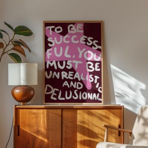 Peut inclure: Une affiche murale encadrée rectangulaire présentant un fond bordeaux profond avec une grande forme d'étoile rose stylisée. Le texte est écrit en lettres blanches style peinture à la main : TO BE SUCCESSFUL, YOU MUST BE UNREALISTIC AND DELUSIONAL.