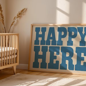 Op de afbeelding: Een ingelijst print met de tekst "HAPPY HERE" in grote, vetgedrukte, blauwe letters op een crèmekleurige achtergrond. De print hangt aan een muur in een kamer met een houten babybedje.