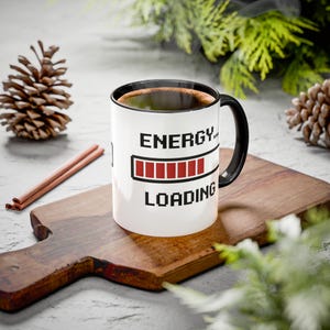 May include: A white ceramic mug with a black handle and interior rim. The exterior features a black pixelated graphic of a loading bar with red segments and the text "ENERGY... LOADING" printed in a bold, black, pixelated font style.
