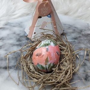 May include: An Easter egg with a floral design in a nest of straw, with a decorative pyramid-shaped gift box tied with a pink ribbon. The gift box has a tag that says "with love".