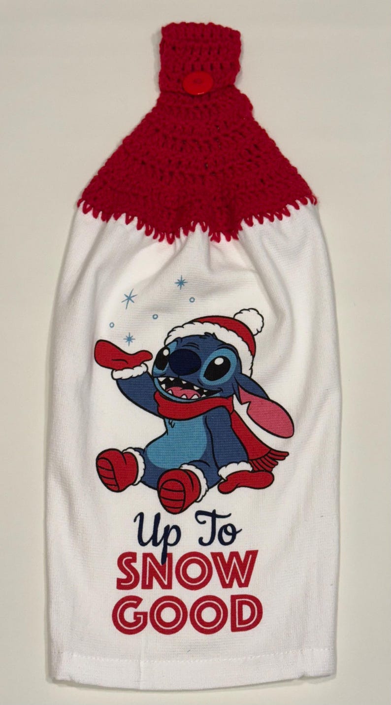 Puede incluir: Toalla de cocina blanca con parte superior de ganchillo roja y cierre de bot&oacute;n. Presenta un personaje de dibujos animados azul con gorro y bufanda de Pap&aacute; Noel, con el texto "Up To SNOW GOOD" en rojo.