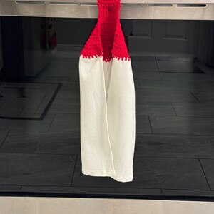 Puede incluir: Una toalla de cocina blanca con una parte superior roja de ganchillo, colgada de un asa de horno de acero inoxidable. La toalla tiene un borde festoneado donde el ganchillo rojo se encuentra con la tela blanca. La toalla de cocina cuelga frente a una superficie de baldosas negras.