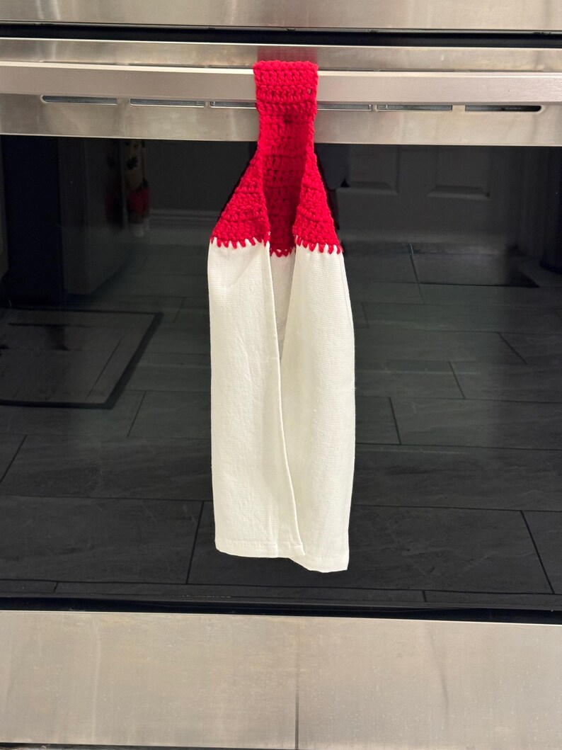 Puede incluir: Una toalla de cocina blanca con una parte superior roja de ganchillo, colgada de un asa de horno de acero inoxidable. La toalla tiene un borde festoneado donde el ganchillo rojo se encuentra con la tela blanca. La toalla de cocina cuelga frente a una superficie de baldosas negras.
