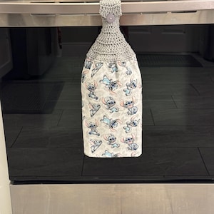 May include: A gray crocheted kitchen towel with a button closure, hanging from an oven handle. The bottom portion features a white fabric printed with a repeating pattern of the character Stitch from Lilo & Stitch in various poses.