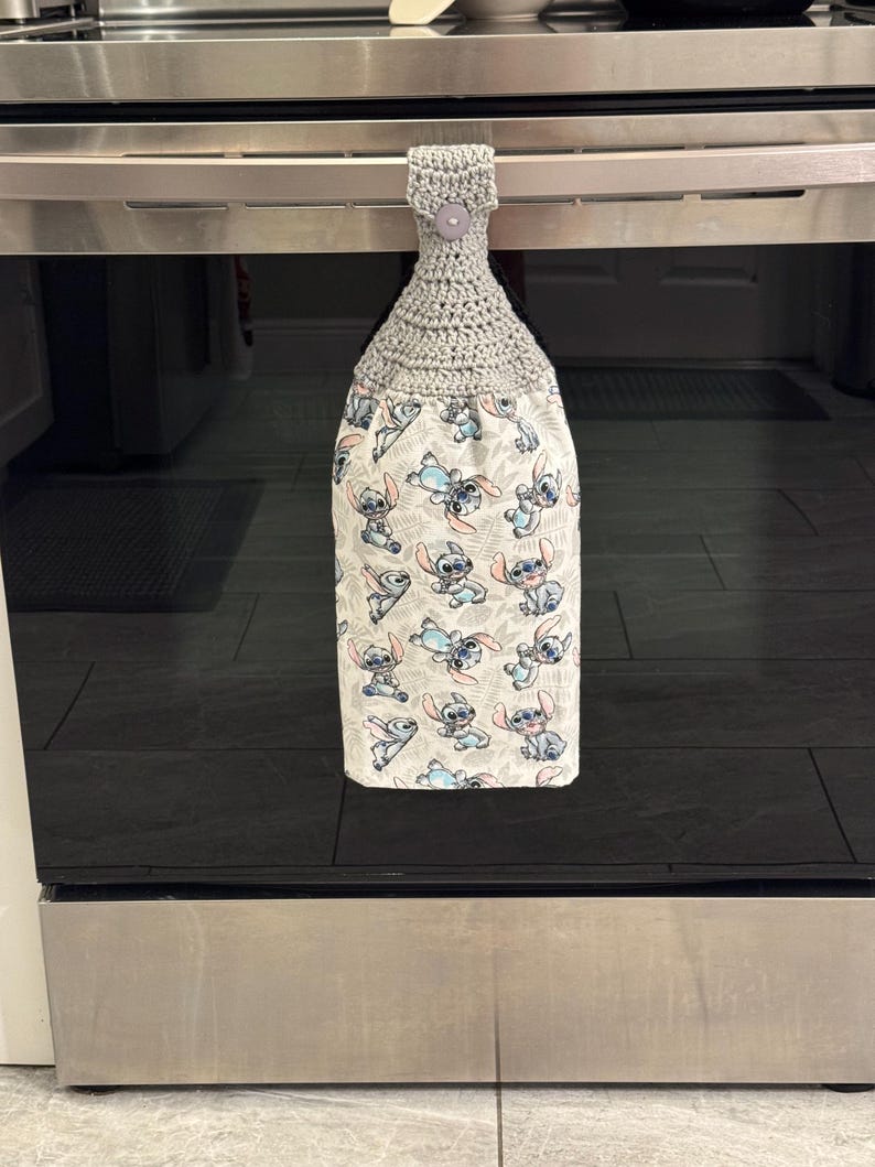 May include: A gray crocheted kitchen towel with a button closure, hanging from an oven handle. The bottom portion features a white fabric printed with a repeating pattern of the character Stitch from Lilo & Stitch in various poses.