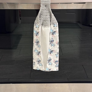May include: A gray crocheted kitchen towel with a loop for hanging. The towel features a white background with a repeating pattern of a blue cartoon character. The towel is hanging on a stainless steel oven.