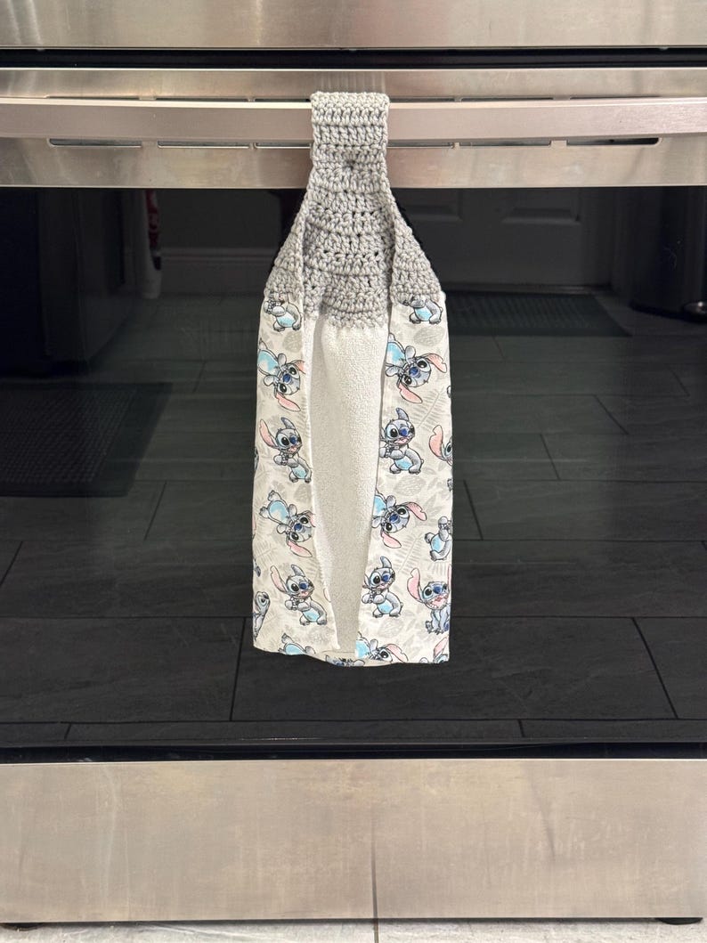 May include: A gray crocheted kitchen towel with a loop for hanging. The towel features a white background with a repeating pattern of a blue cartoon character. The towel is hanging on a stainless steel oven.