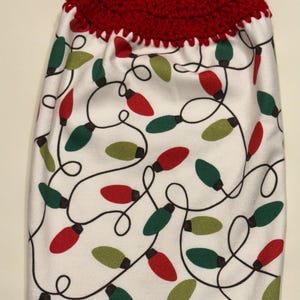 May include: A white kitchen towel with a red crocheted top and button fastening. The towel has a pattern of red and green Christmas lights with black wiring.