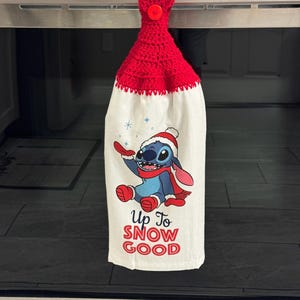 Puede incluir: Un pa&ntilde;o de cocina de ganchillo rojo con cierre de bot&oacute;n. La tela blanca presenta un personaje de dibujos animados azul con gorro y bufanda de Pap&aacute; Noel, con el texto "Up To SNOW GOOD" en rojo.