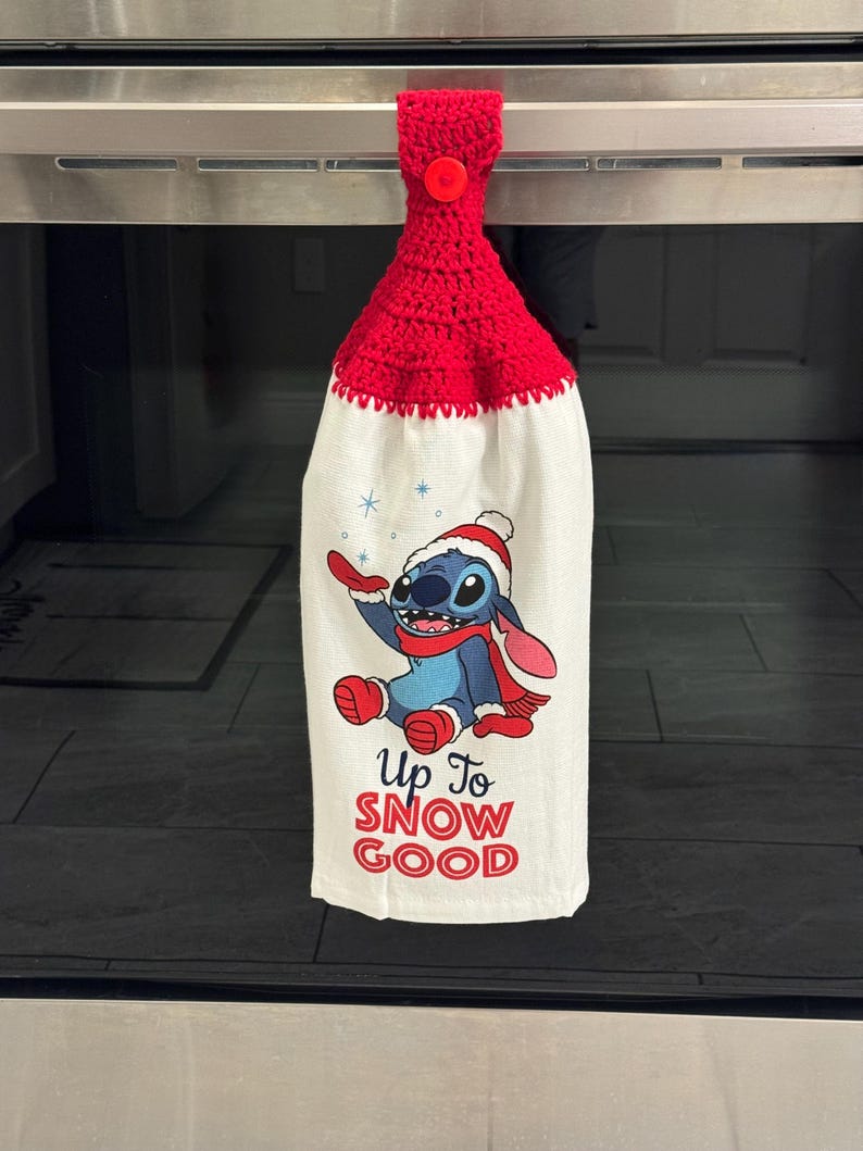 Puede incluir: Un pa&ntilde;o de cocina de ganchillo rojo con cierre de bot&oacute;n. La tela blanca presenta un personaje de dibujos animados azul con gorro y bufanda de Pap&aacute; Noel, con el texto "Up To SNOW GOOD" en rojo.