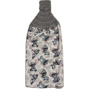 May include: A hanging kitchen towel featuring a gray crocheted top with a button closure. The towel's fabric is white with a repeating pattern of the character Stitch from the movie Lilo & Stitch, in shades of blue and pink.