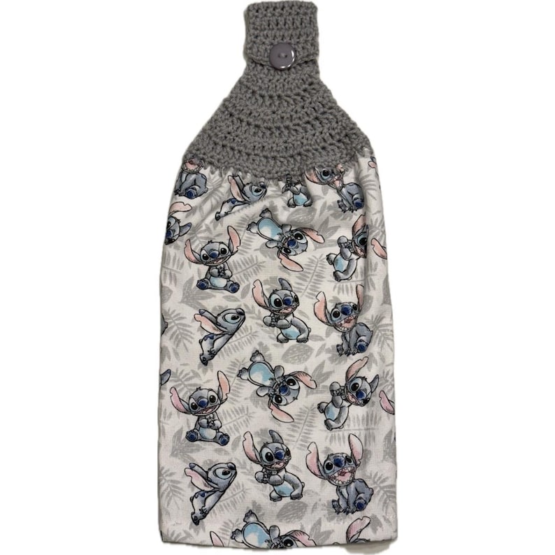 May include: A hanging kitchen towel featuring a gray crocheted top with a button closure. The towel's fabric is white with a repeating pattern of the character Stitch from the movie Lilo & Stitch, in shades of blue and pink.