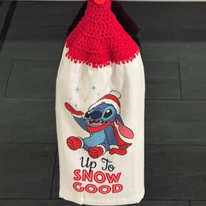 Puede incluir: Un pa&ntilde;o de cocina blanco con una parte superior y un bot&oacute;n de ganchillo rojos. La toalla presenta un personaje de dibujos animados con gorro y bufanda de invierno, con el texto "Up To SNOW GOOD" en rojo.